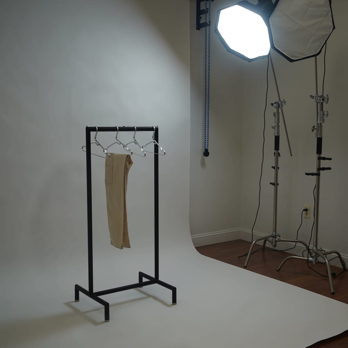 A clothing rack holding a pair of beige pants stands in a photography studio with a white backdrop and lighting equipment.
