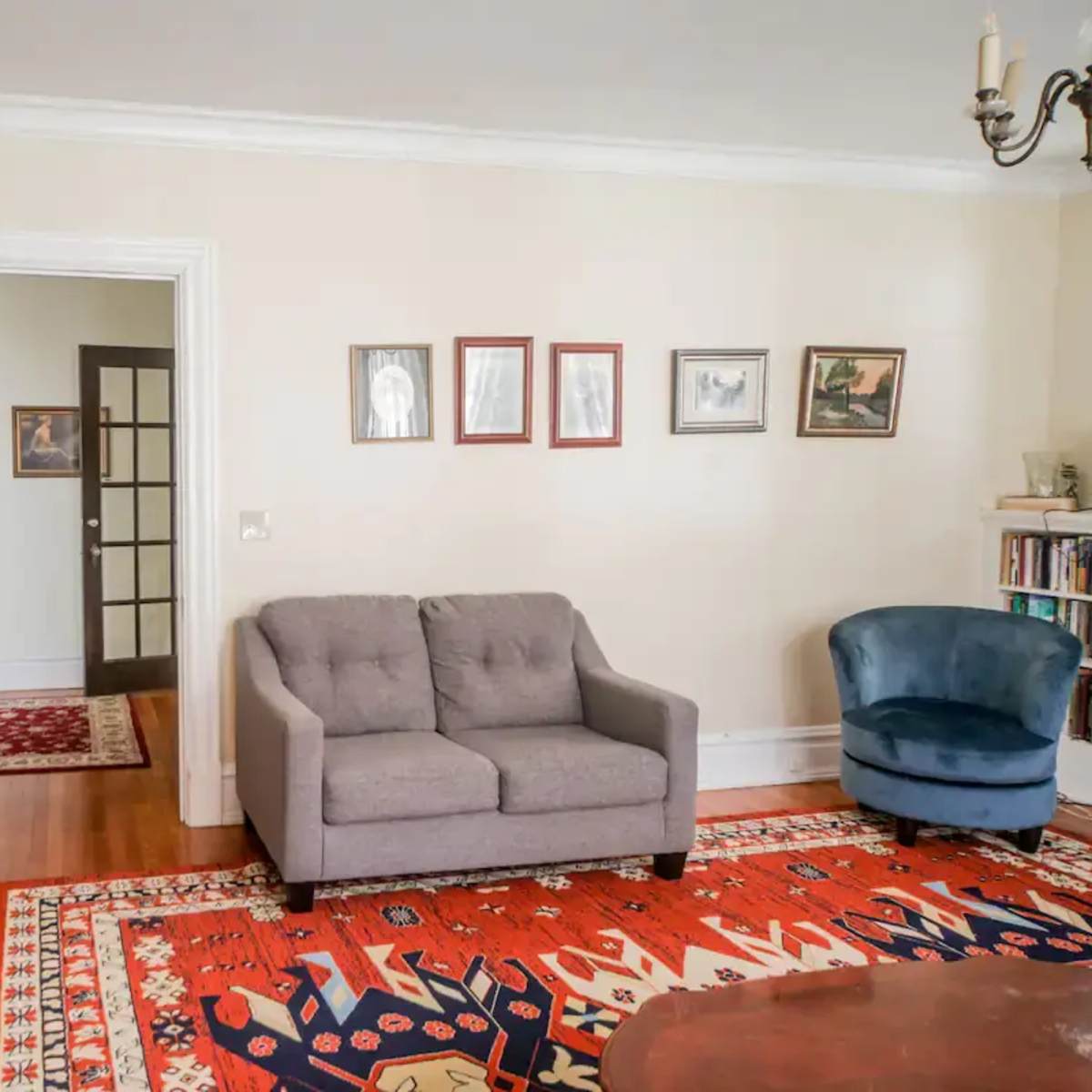 The room features a gray sofa and a blue armchair, with framed artwork on the wall opposite a wooden furniture piece and a doorway leading to another area.