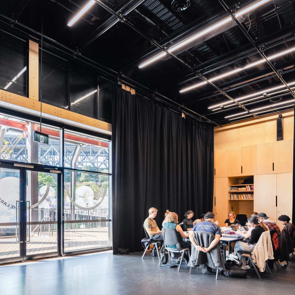 A group of people sits around a table in a well-lit room with large windows and black curtains, overlooking an industrial landscape.