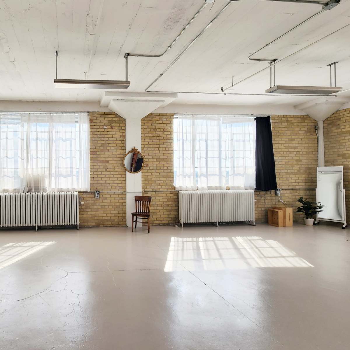 A spacious room with exposed brick walls, large windows, and minimal decor features a chair, a mirror, and a potted plant.
