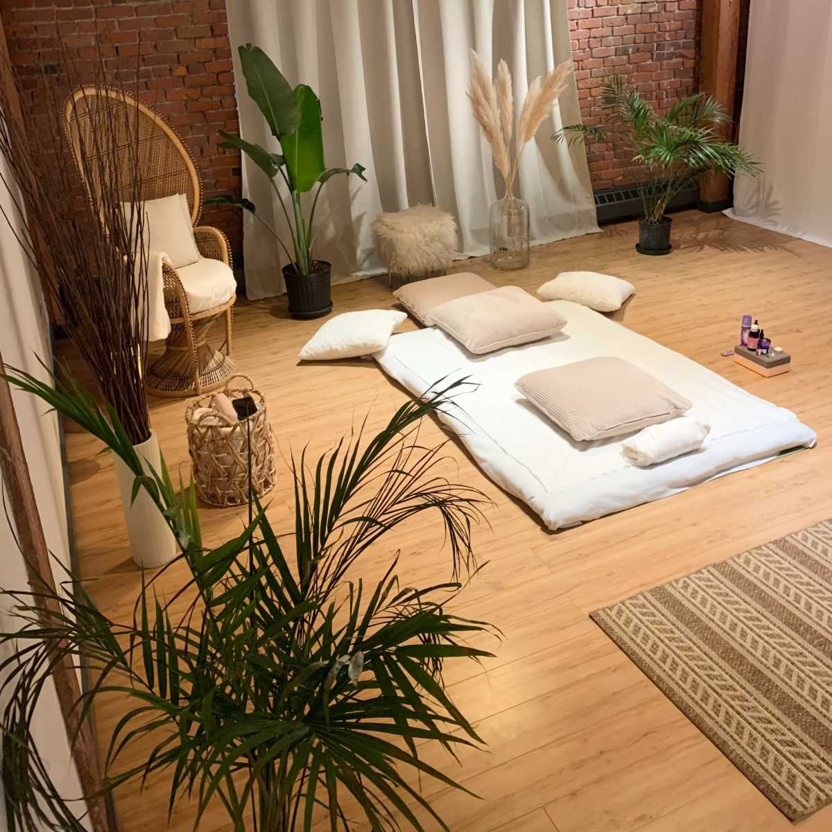 The scene features a cozy minimalist space with a large white mat on the wooden floor, surrounded by plants and soft cushions, and a backdrop of draped fabric and exposed brick.