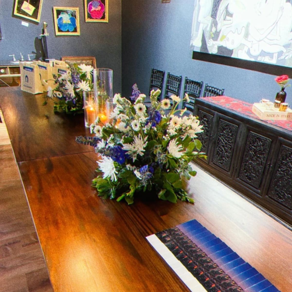 A wooden table adorned with floral centerpieces and lit candles, accompanied by boxes and a wall featuring framed artwork.