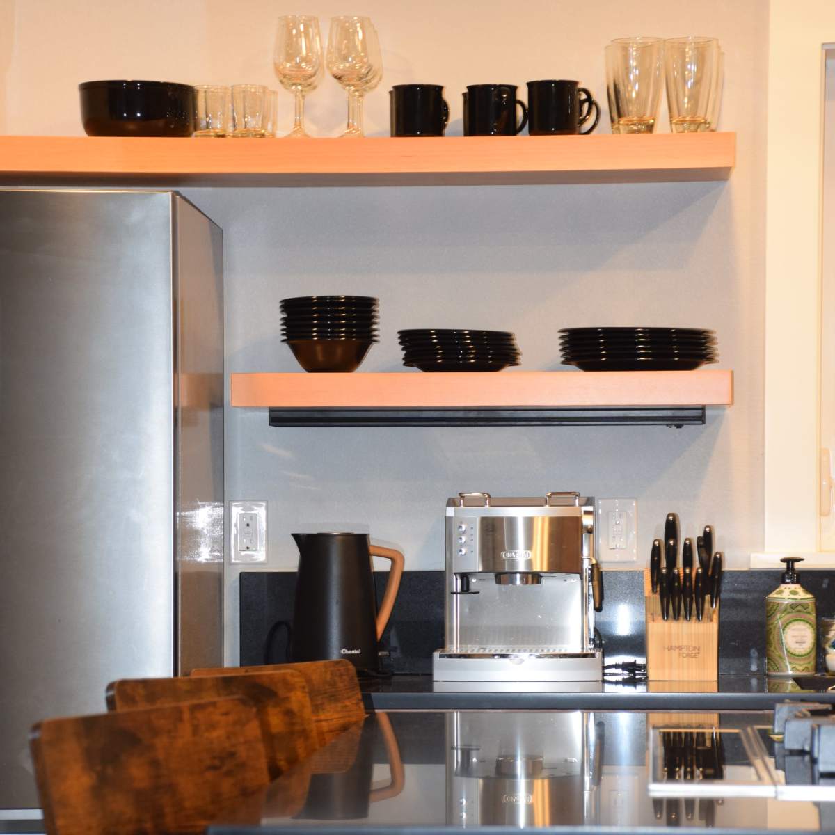 A modern kitchen features stainless steel appliances, wooden bar stools, and neatly arranged glassware and dishes on shelves.