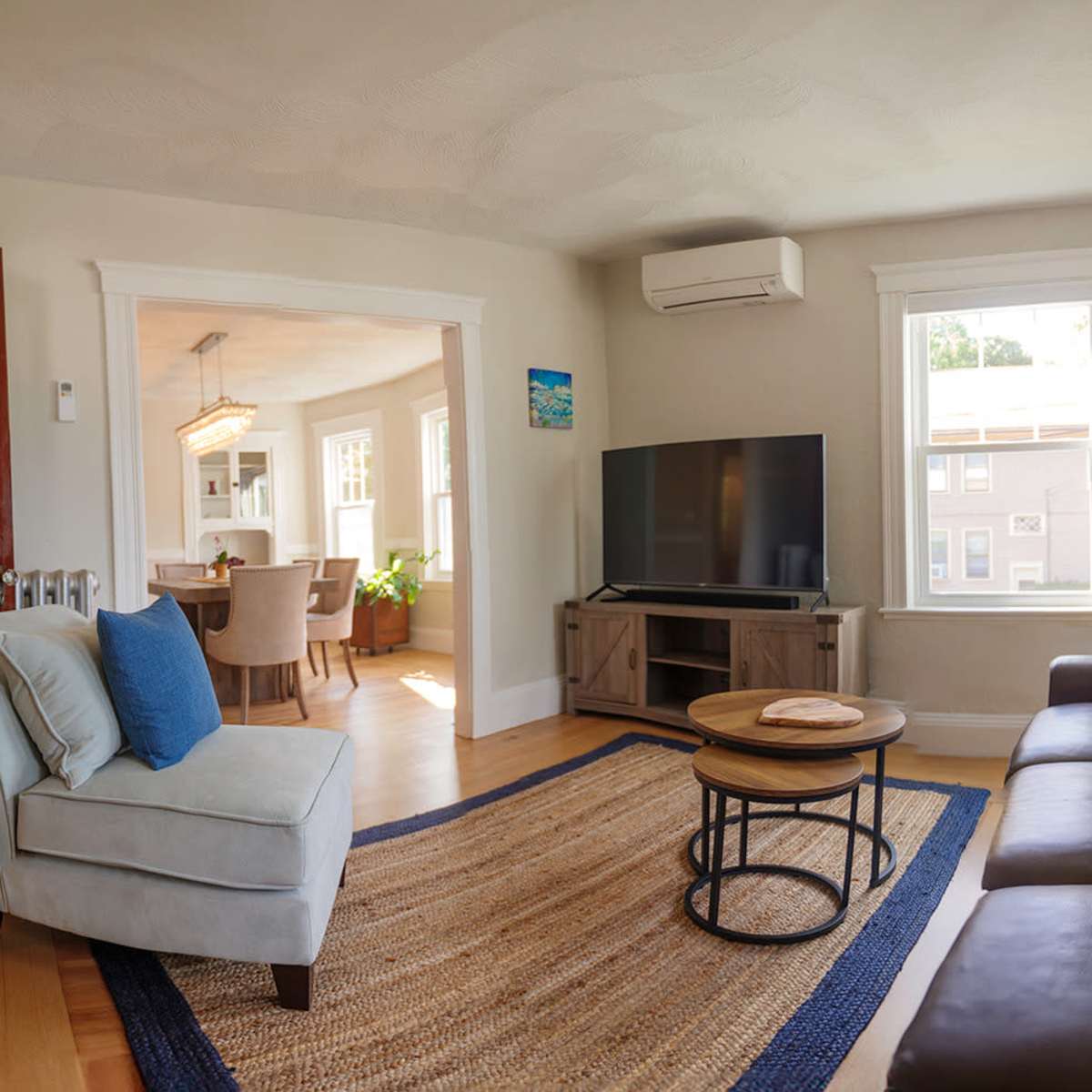 A living room features a brown sofa, a light blue chair, a round coffee table, and a television, with windows allowing natural light and a view into a dining area.