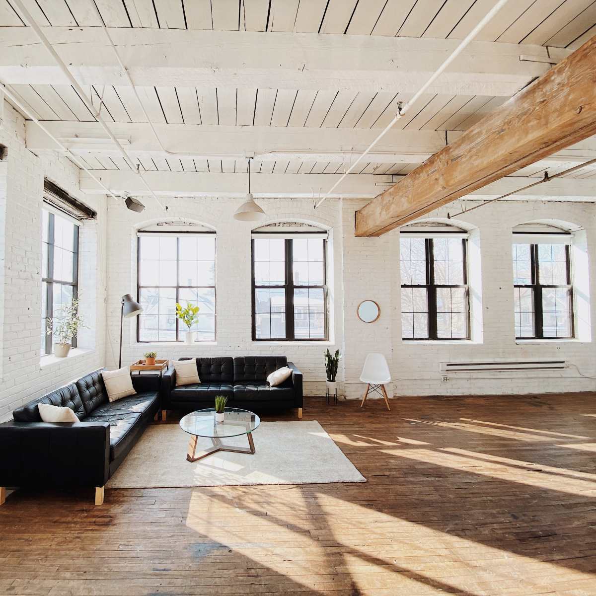 A spacious, sunlit loft features a black sectional sofa, a glass coffee table, and several plants arranged on window sills.