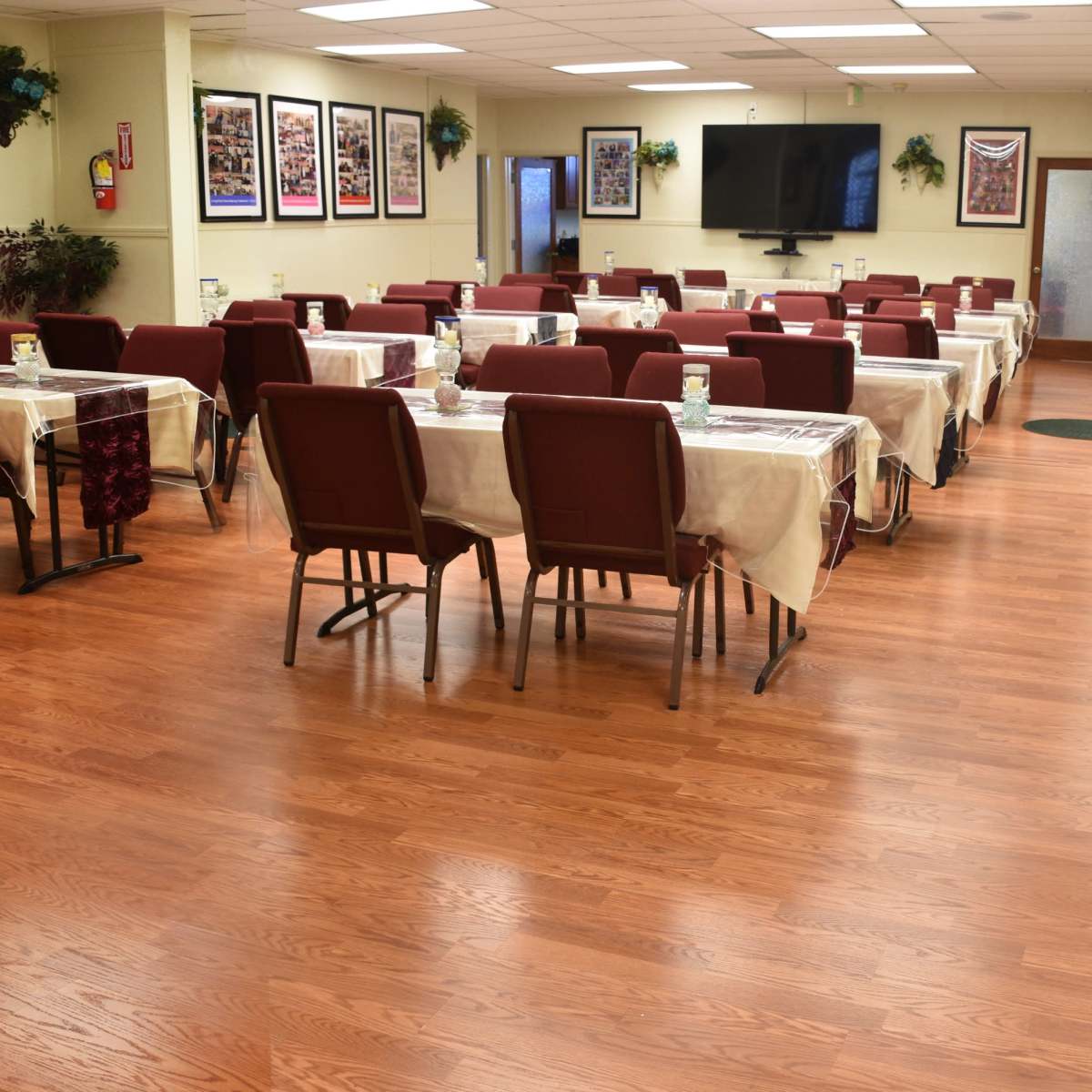 A spacious dining area features rows of empty tables covered with white tablecloths and surrounded by red chairs, with framed photographs on the walls.