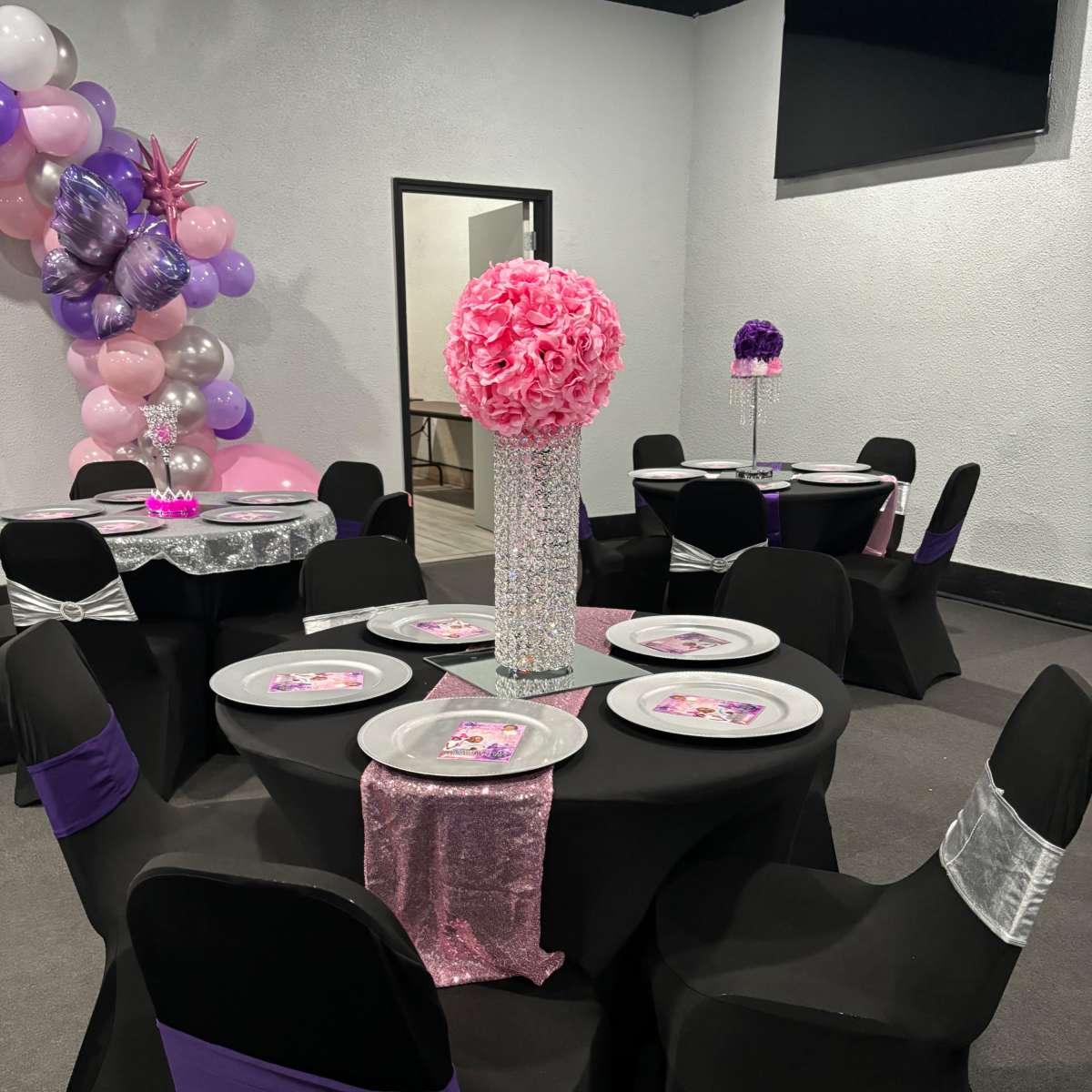 A decorated event space with black tablecloths, chairs covered in black with purple and silver accents, and floral centerpieces.