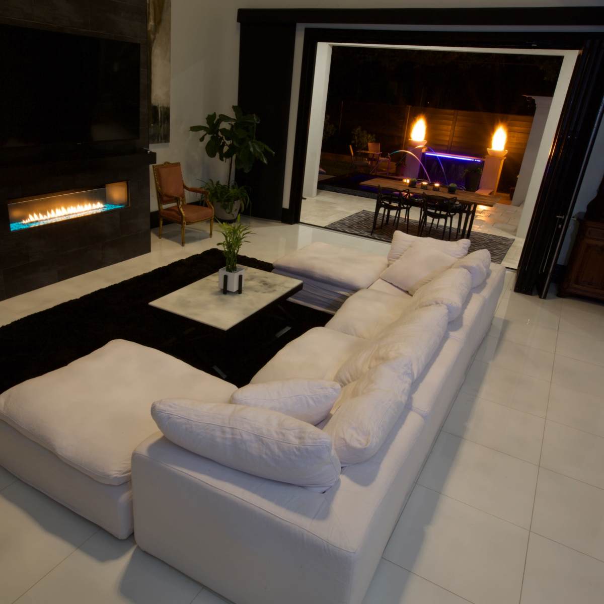 A modern living room with a large white sectional sofa, a coffee table, and a sleek fireplace, leading to an outdoor area with illuminated elements.