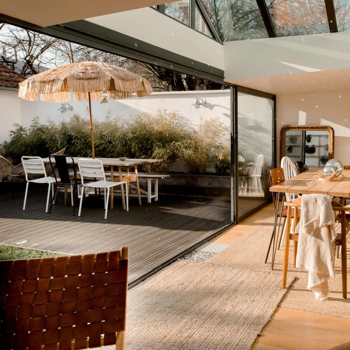 A sunlit dining area features a wooden table surrounded by chairs, with a glass wall opening to an outdoor space that includes a table with an umbrella.