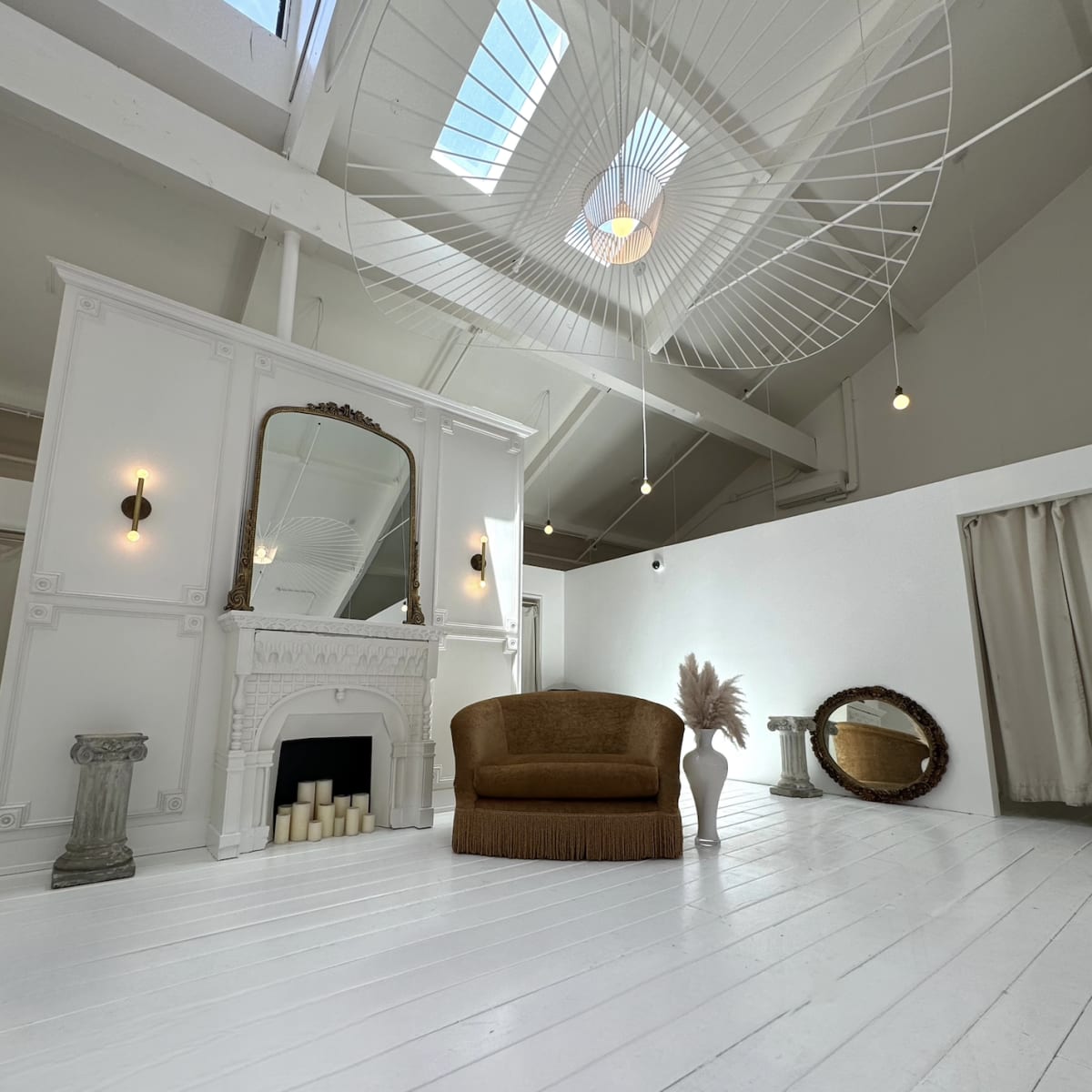 A spacious, bright room with white wooden floors, a large mirror, a brown couch, and decorative elements like a fireplace and vases.