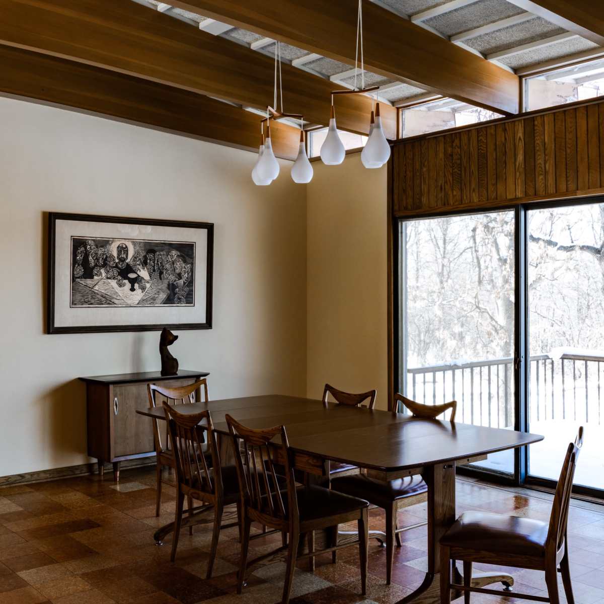 A modern dining room with wooden beams, a large table surrounded by chairs, and a framed artwork on the wall near a window with views of a snowy landscape outside.