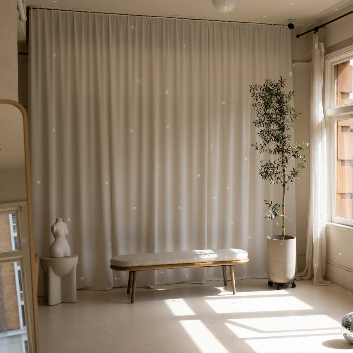 A sunlit room features a bench, a potted plant, floor-to-ceiling curtains, and a reflective sphere on the floor.