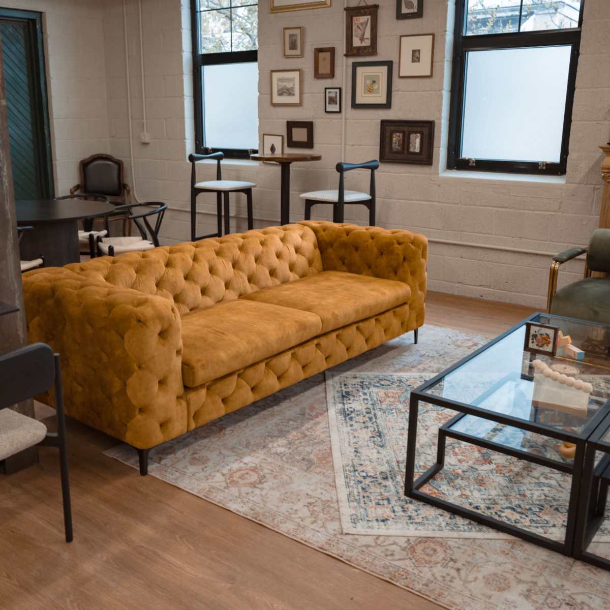 A cozy interior with a mustard-colored tufted sofa, a glass coffee table, and various framed pictures on the walls.