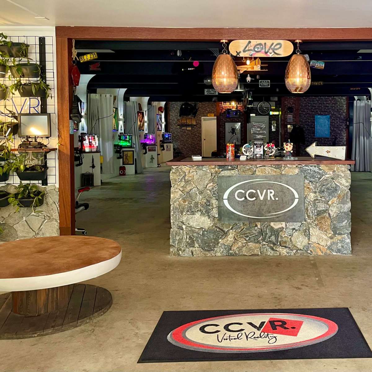 A modern reception area for a virtual reality venue, featuring a stone counter, potted plants, and gaming stations in the background.