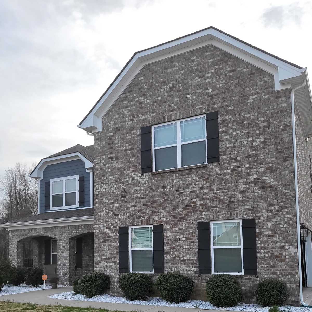 A two-story brick house with a combination of brown and blue siding, featuring black shutters and landscaped shrubbery in front.
