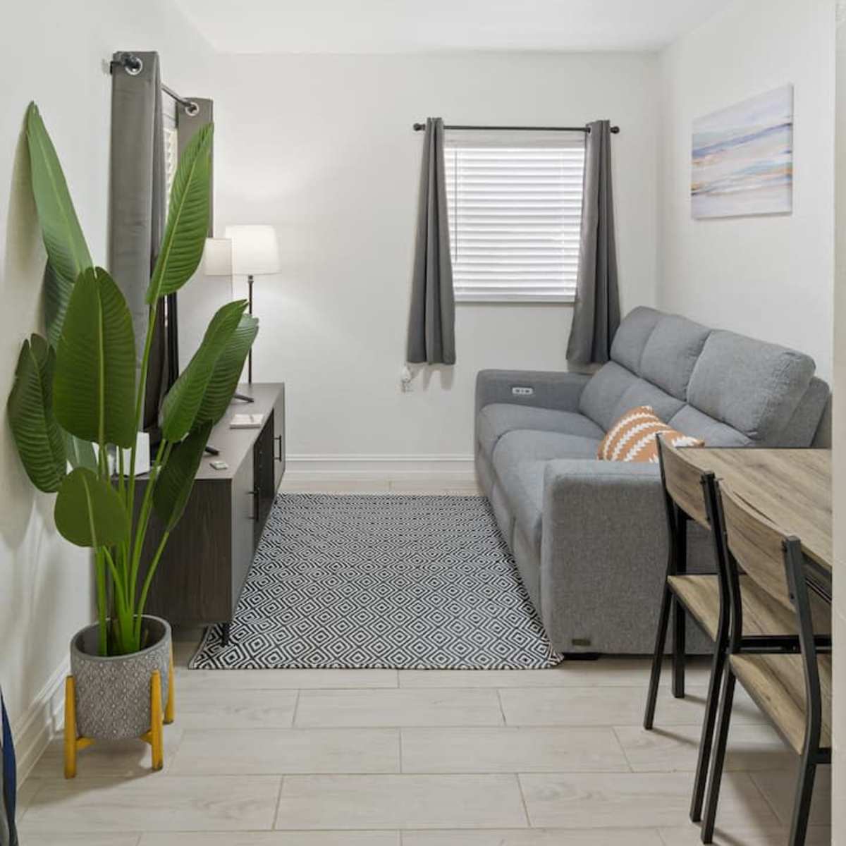 A modern living room with a gray sofa, a television stand, a small dining area, and a large indoor plant.