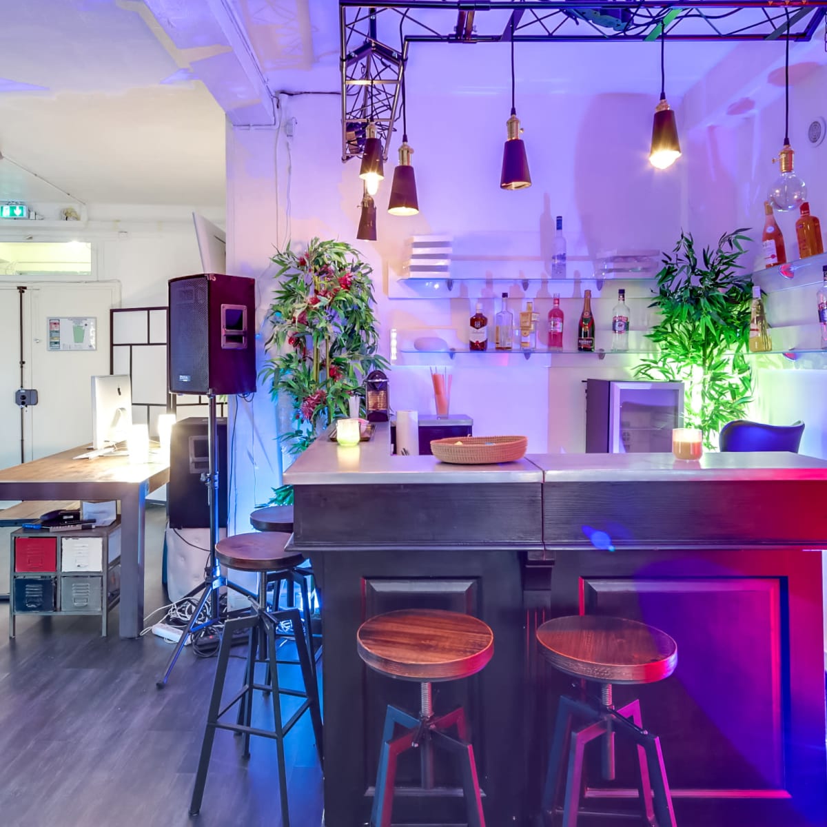 A modern bar area with three wooden stools, a sleek counter, colorful lighting, and shelves displaying various bottles of liquor.