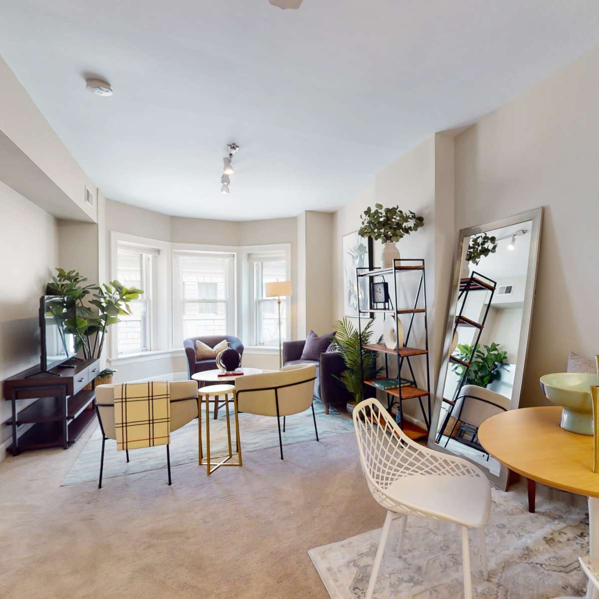 A well-furnished living space featuring a seating area with a round table, a television stand, and large windows with natural light, surrounded by decorative plants and a mirror.