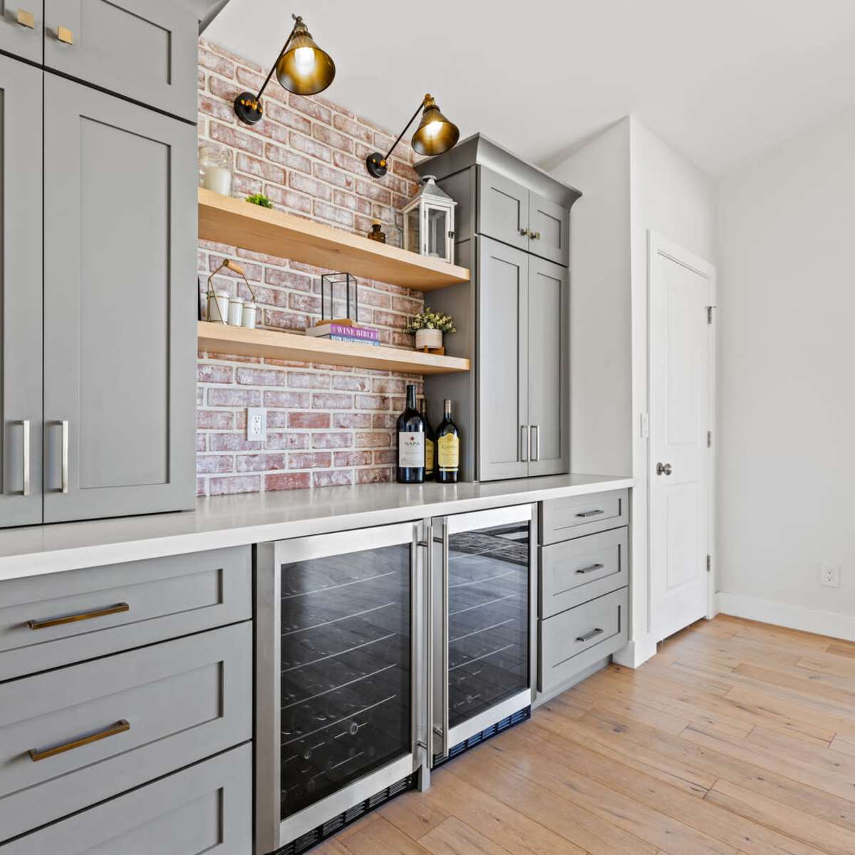 A modern kitchen bar area with gray cabinets, a white countertop, and open wooden shelves above a brick accent wall.