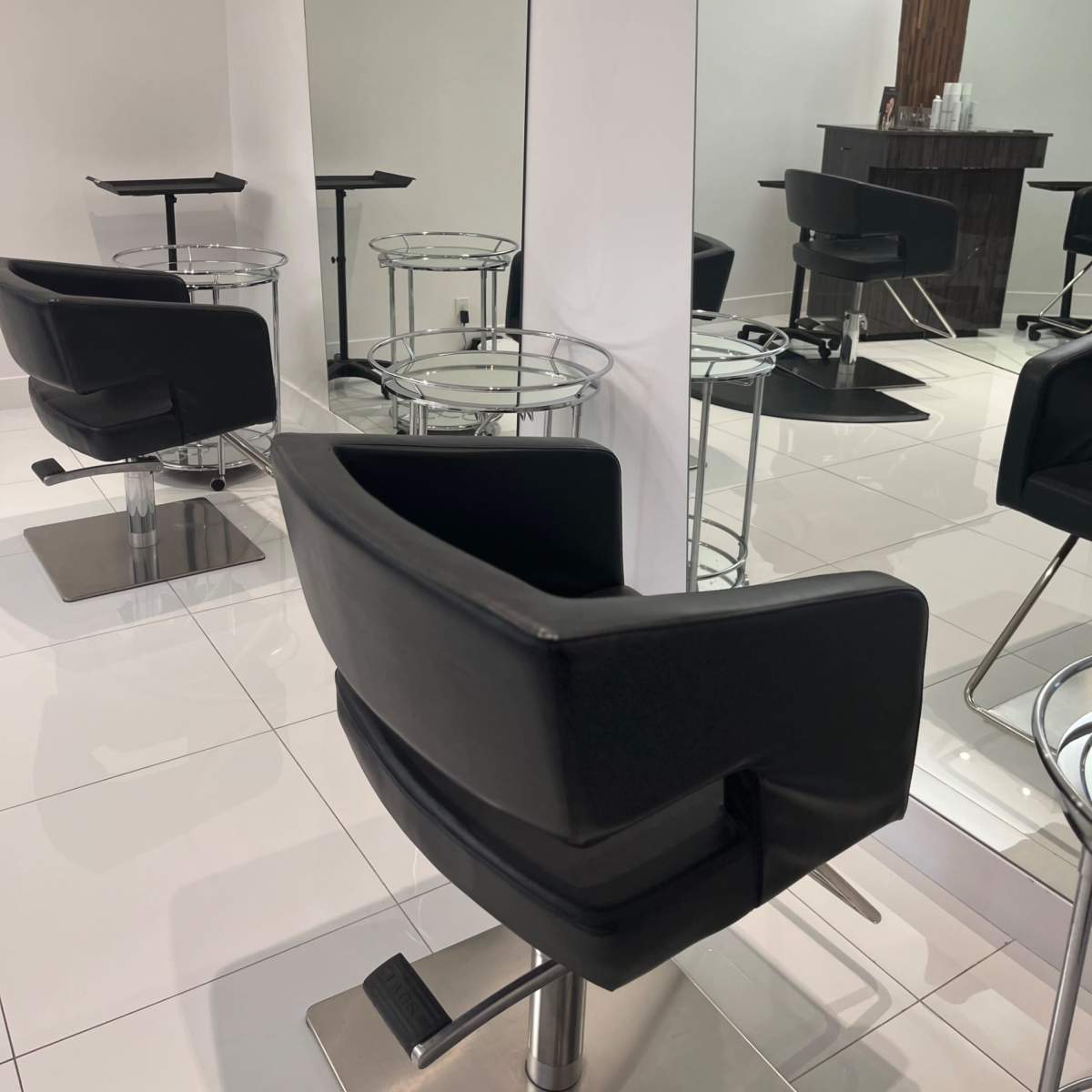 A modern salon interior with black reclining chairs and mirrors arranged against white walls and tiled floors.