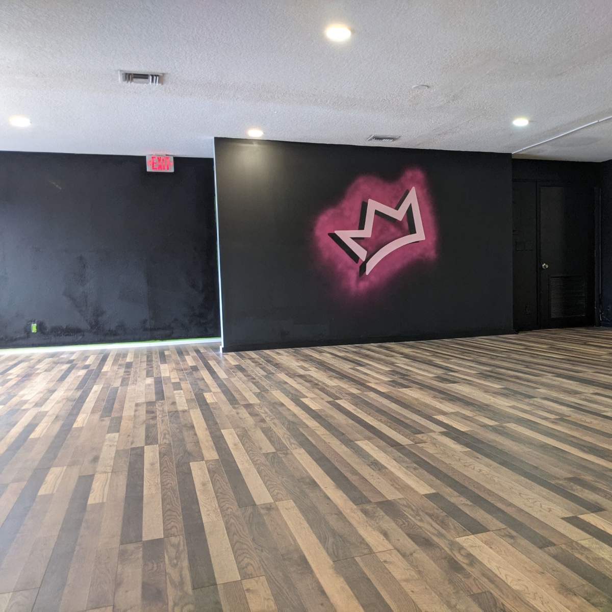 An empty studio space with wooden flooring and a black wall featuring a pink crown graphic.