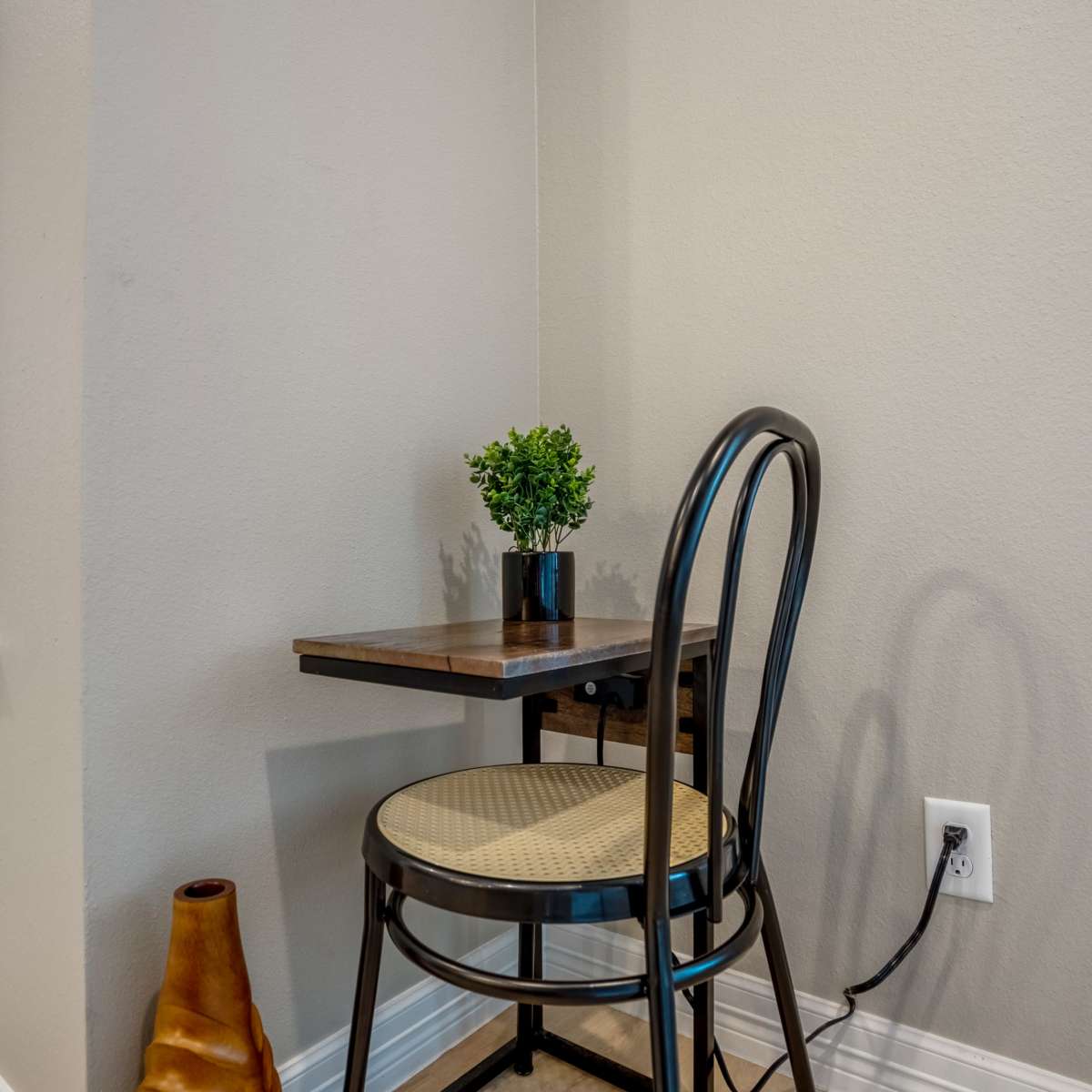 A small seating area features a wooden table in the corner with a chair and a potted plant on top, next to a wall with a power outlet.