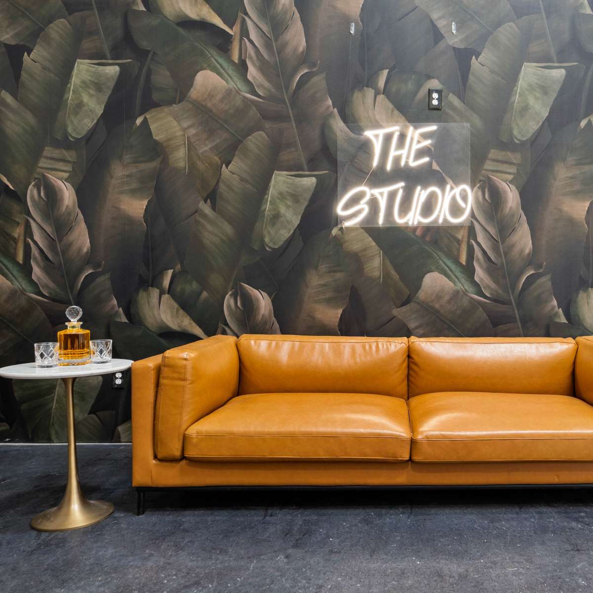 A tan leather sofa is positioned in front of a leafy wallpaper, with a neon sign reading "THE STUDIO" and a vintage lamp beside a glass table with a decanter.