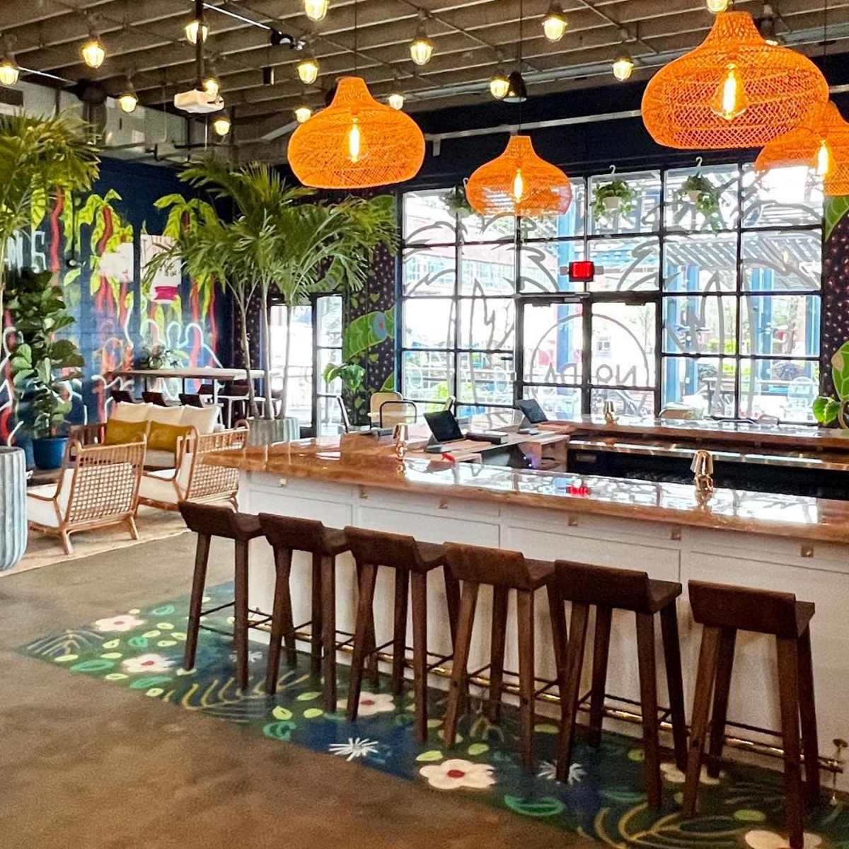 A stylish indoor space featuring a bar with seating, greenery, and colorful mural art on the walls.