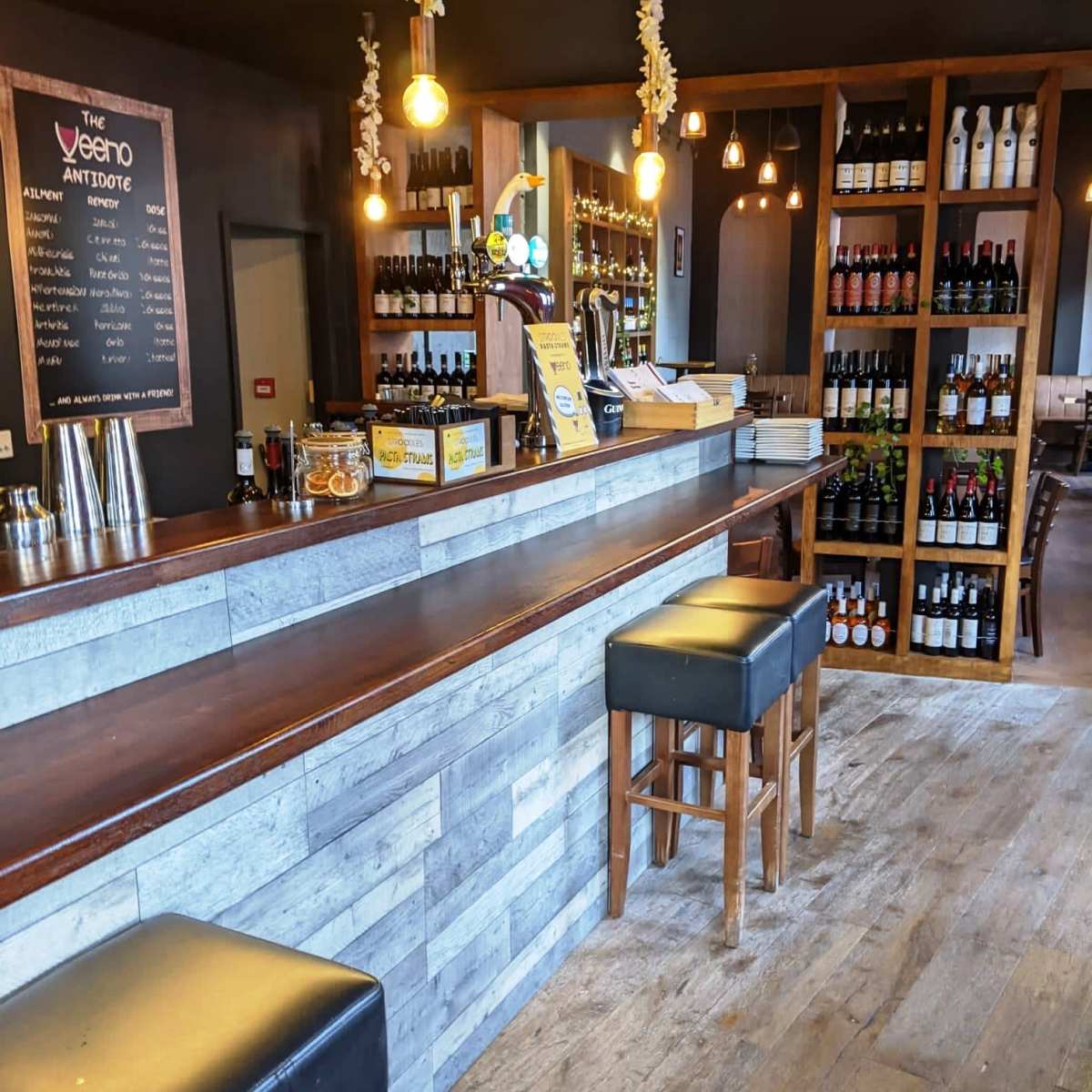 A bar area with a wooden counter, high stools, shelves filled with bottles, and warm lighting.