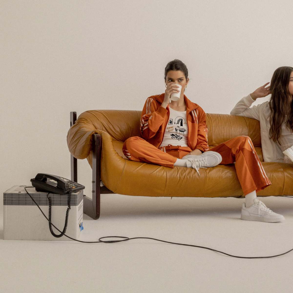 Two young women sit on a mustard-colored couch, with one holding a beverage and the other adjusting her hair, while a vintage telephone sits on a nearby box.