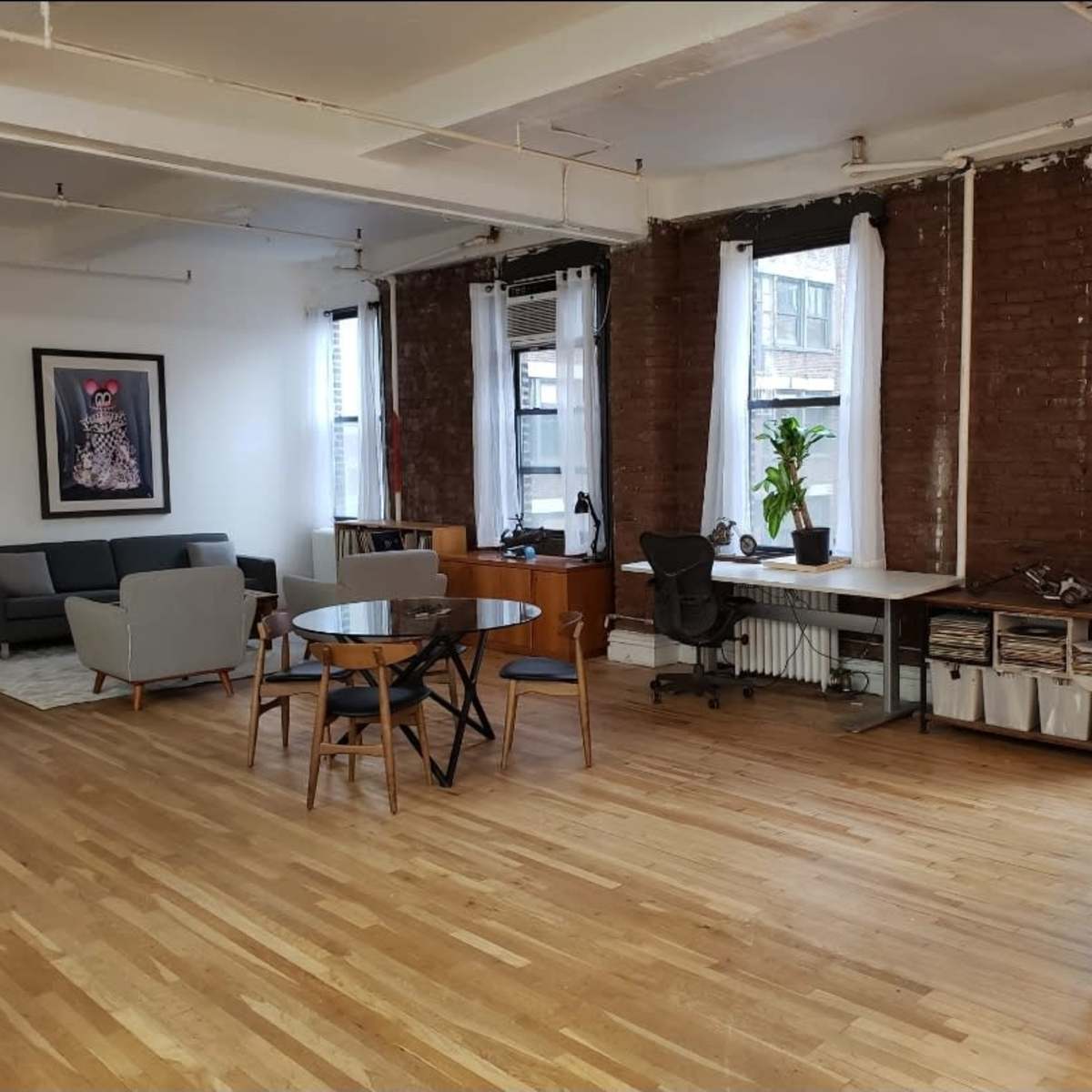 A spacious room with wooden flooring, featuring a sitting area with gray sofas, a dining table with chairs, and a desk against a brick wall with large windows.