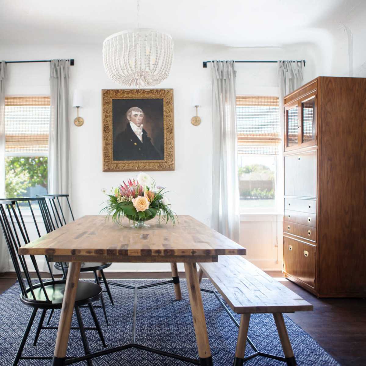 A dining area features a rustic wooden table, black chairs, a floral centerpiece, a chandelier, and a portrait hanging on the wall.