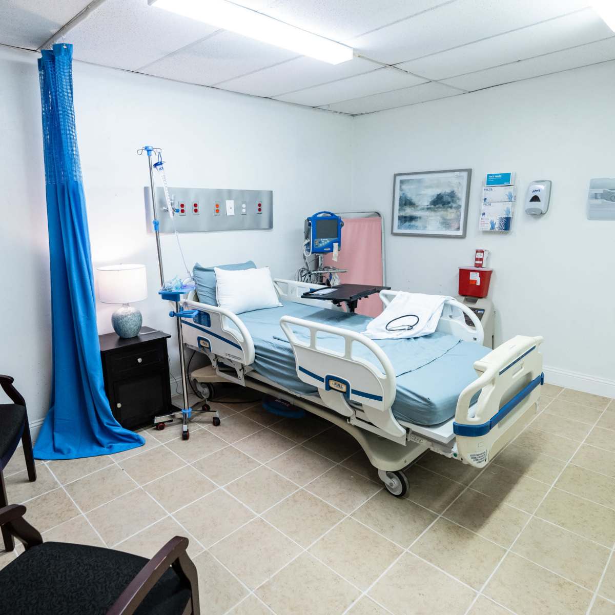 A hospital room featuring a patient bed, medical equipment, a small bedside table, and a chair in a tidy environment.