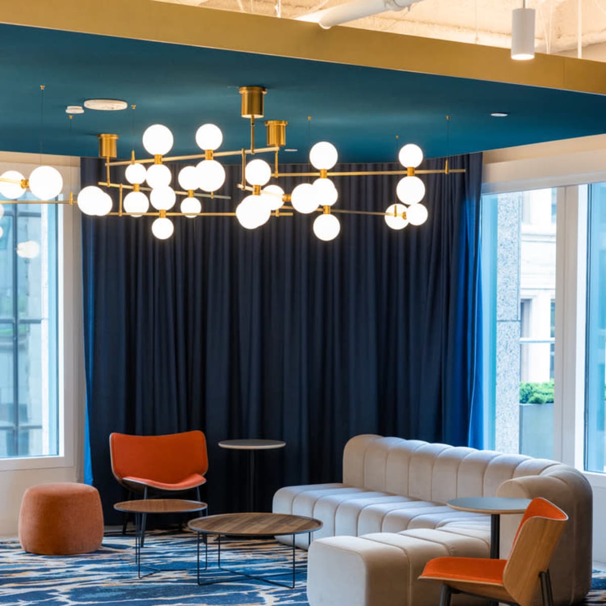 A modern lounge area featuring a gray sofa, orange chairs, a small round table, and a chandelier with multiple light bulbs, set against a backdrop of dark curtains and large windows.