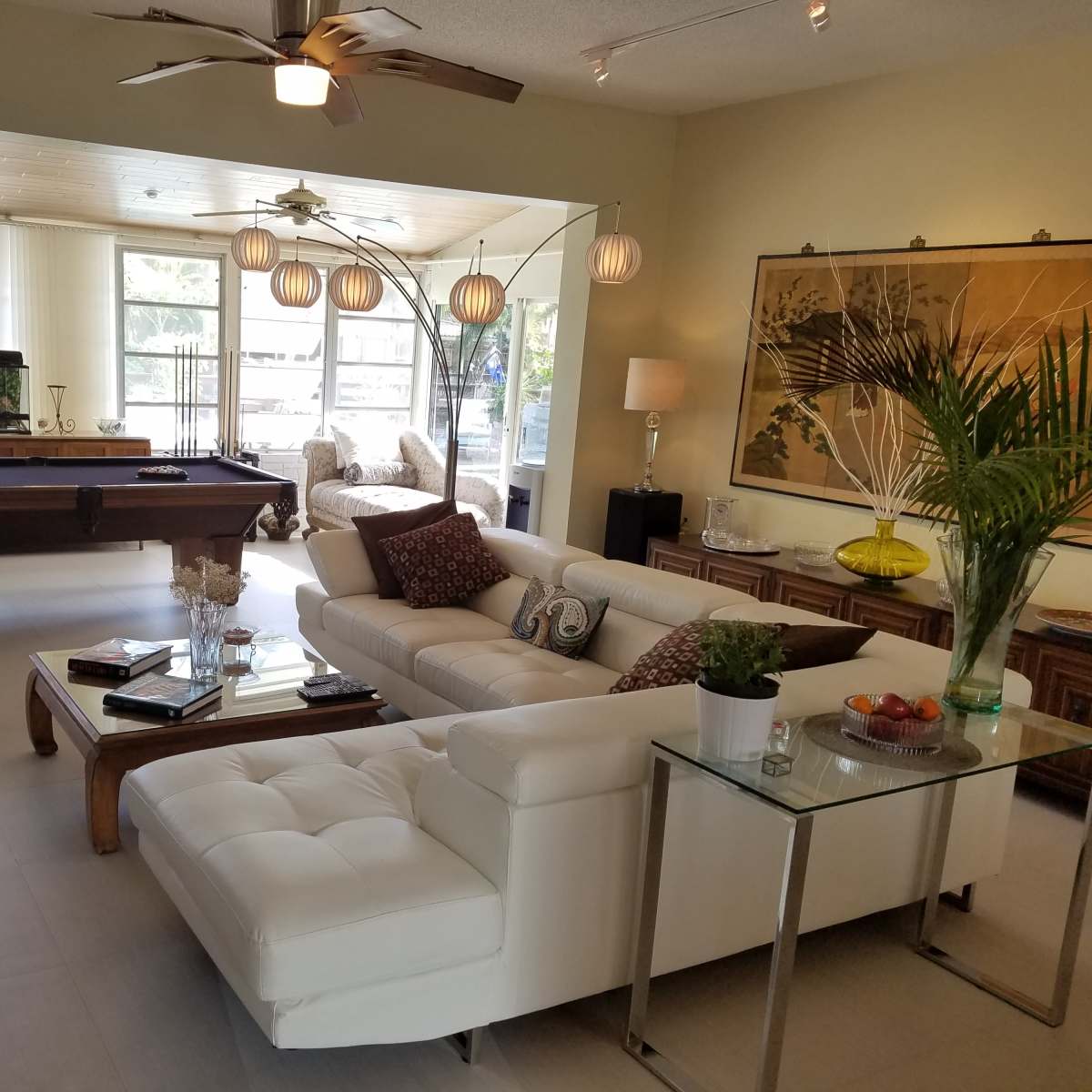 A spacious living room with a white sectional sofa, a pool table, and large windows that provide ample natural light.