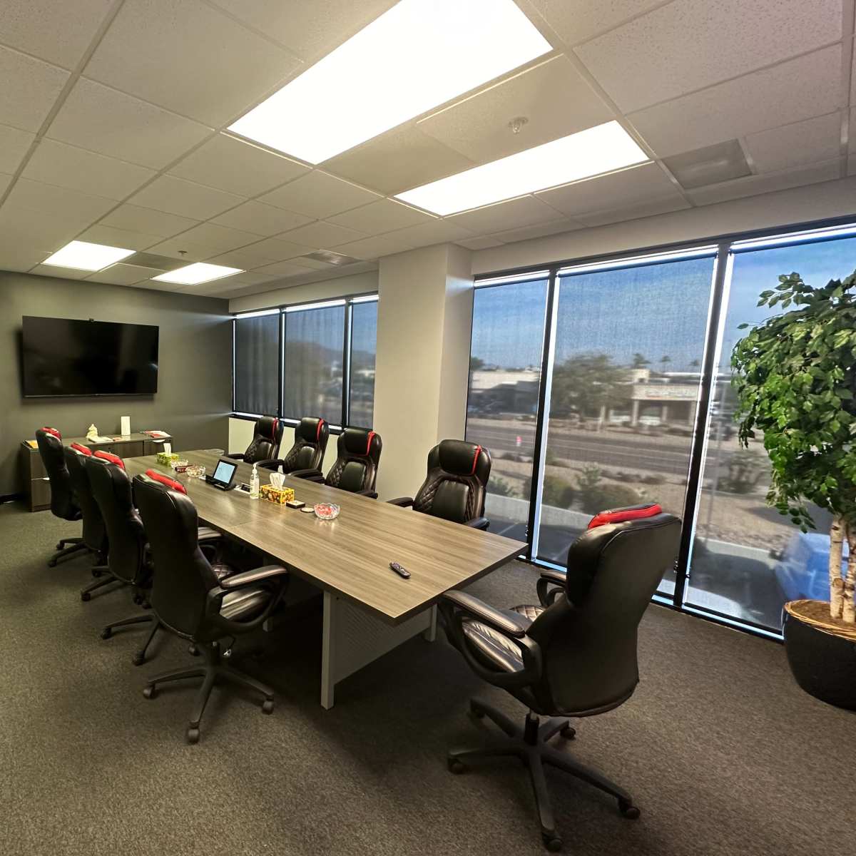 A conference room features a large table surrounded by ergonomic chairs, a wall-mounted TV, and large windows overlooking an outdoor landscape.