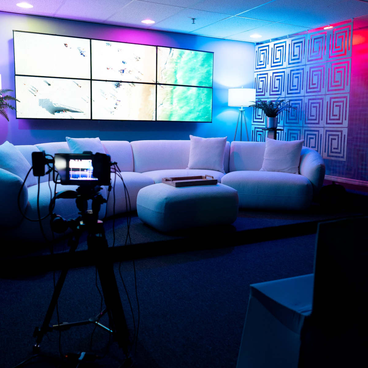 A modern studio setup featuring a white sectional sofa, a large display screen on the wall, and various lighting elements.