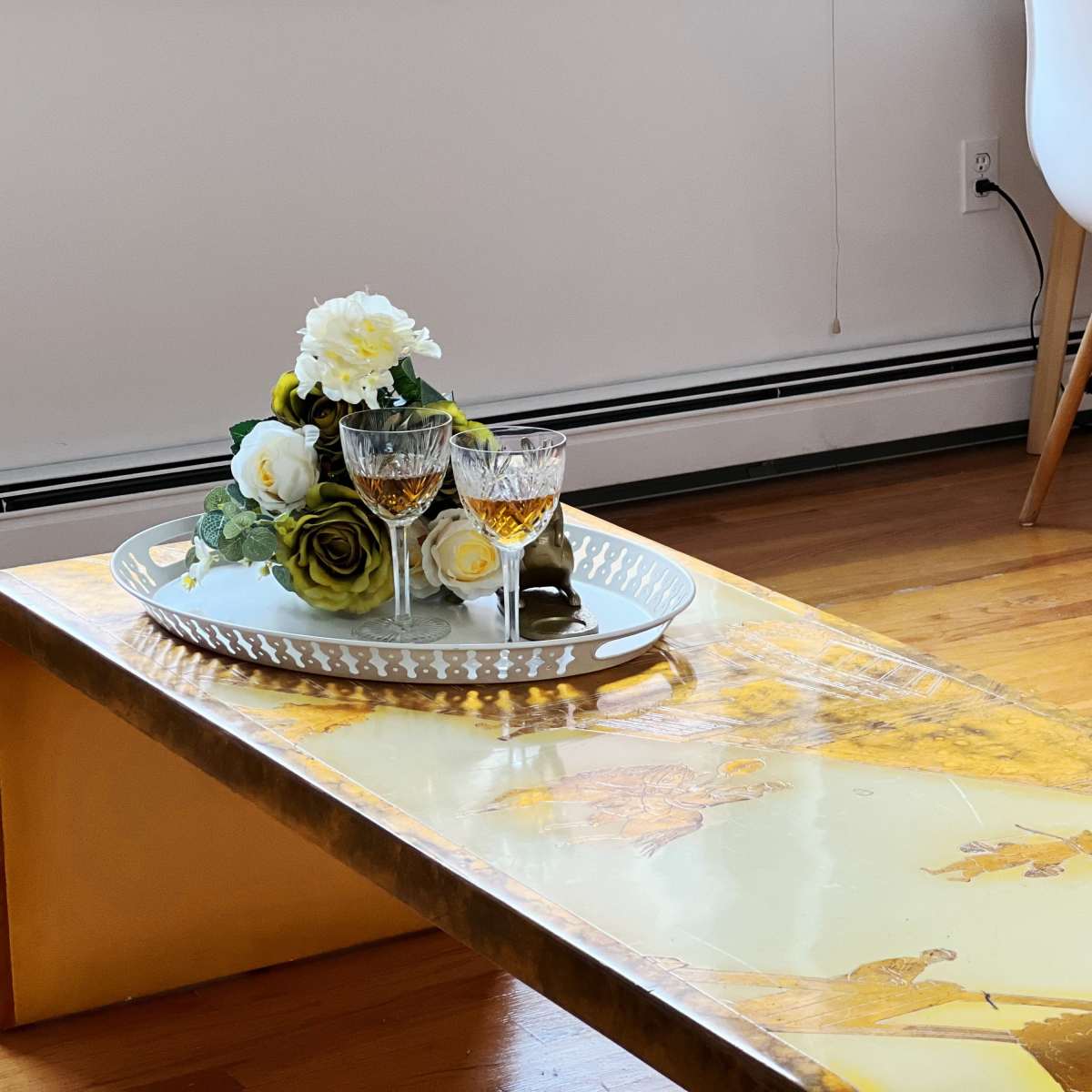 A wooden coffee table displays a decorative tray with two glasses and a floral arrangement, while a white desk is visible in the background.
