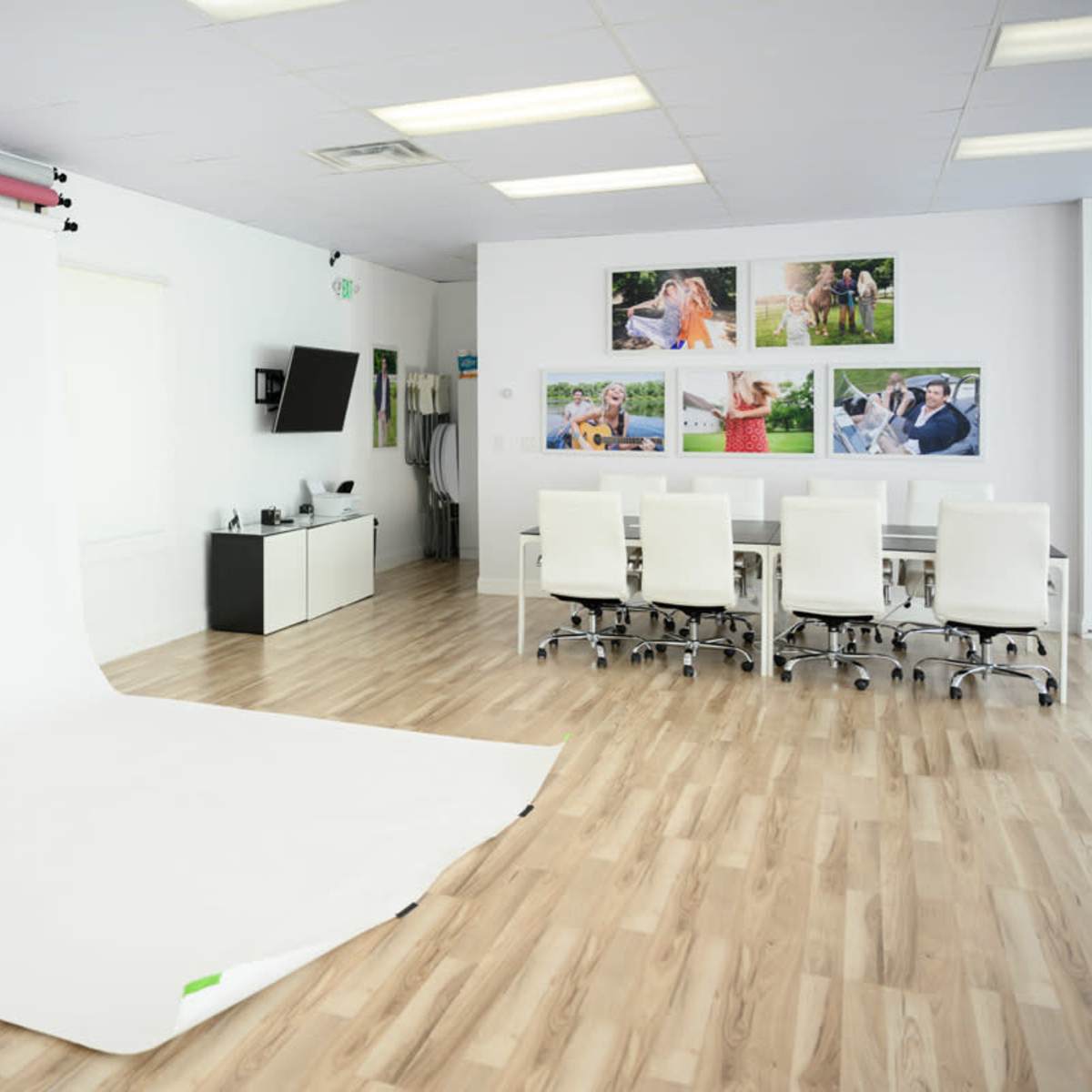 A photography studio with a white backdrop, a table surrounded by white chairs, and framed photographs on the wall.