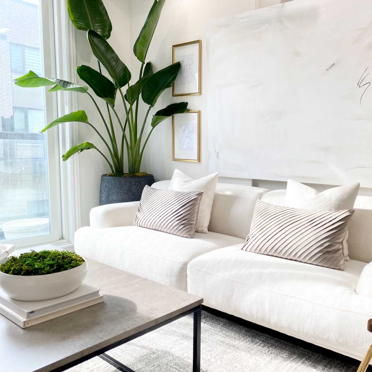 A modern living room features a white sofa with striped pillows, a large abstract painting, and a green indoor plant next to a glass window.