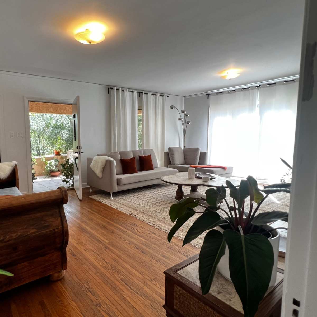 A bright living room featuring a wooden sofa, a coffee table, and two armchairs, with a view of an outdoor area through an open doorway.
