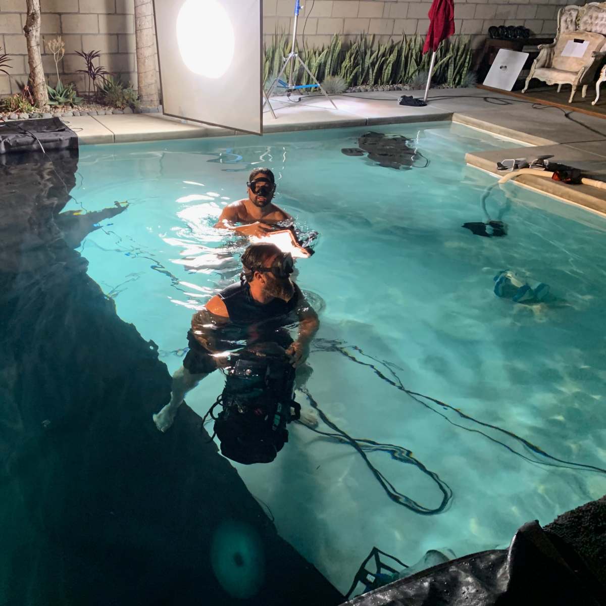 Two individuals are submerged in a pool while filming, with equipment positioned around the water.
