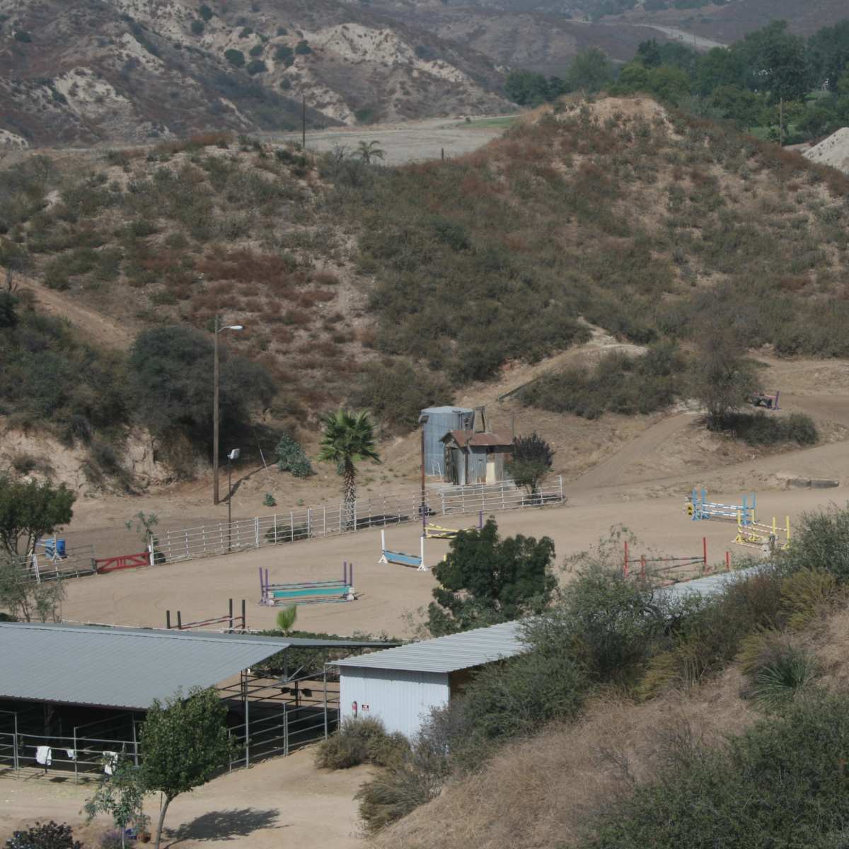 A horse riding arena situated in a dry, hilly landscape, surrounded by sparse vegetation and a few structures.