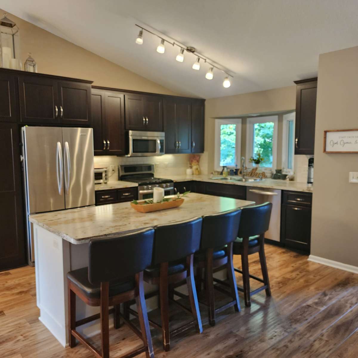 A modern kitchen features dark wood cabinetry, stainless steel appliances, a large island with seating, and natural light streaming through the windows.