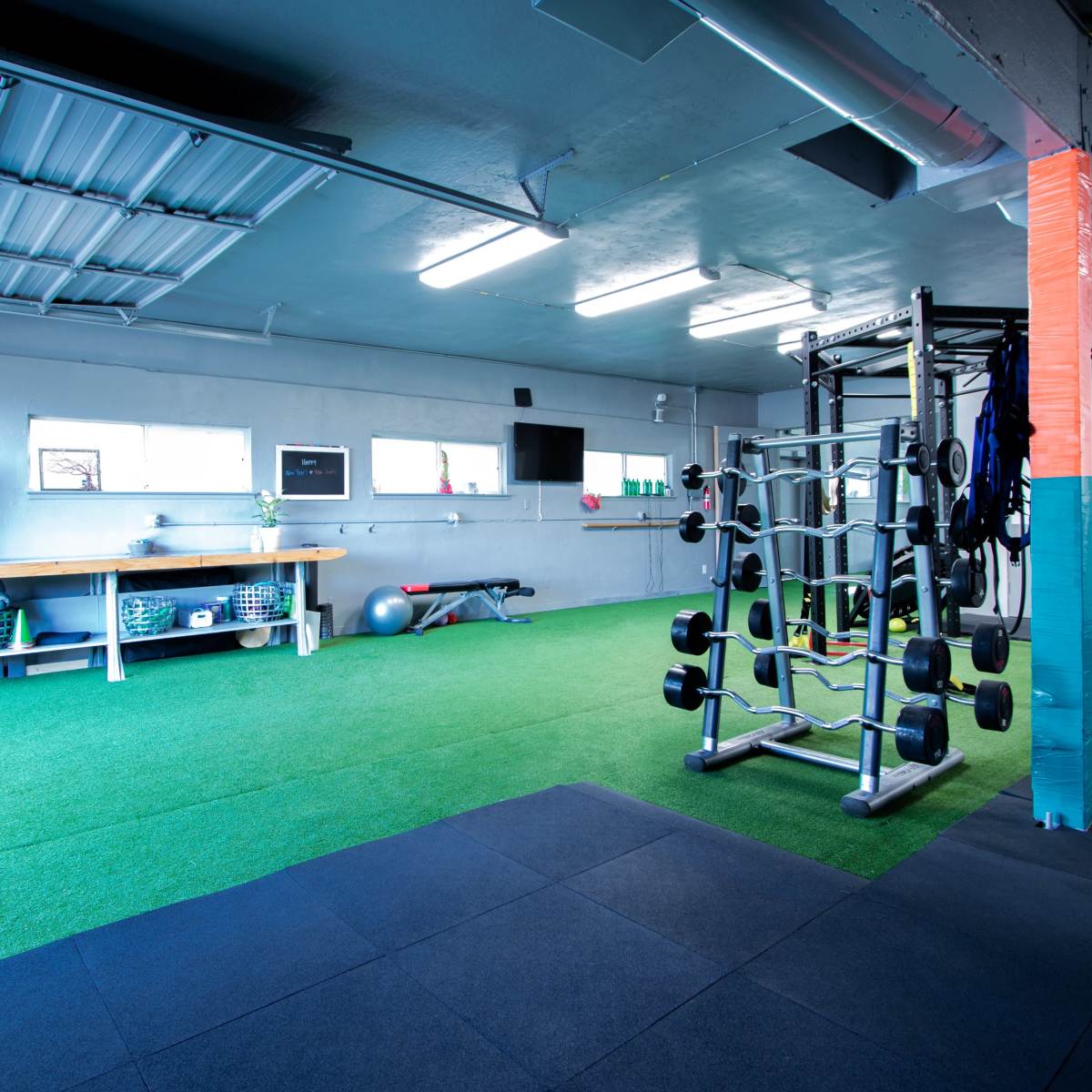A spacious gym with green turf flooring, a weight rack, exercise equipment, and large windows providing natural light.