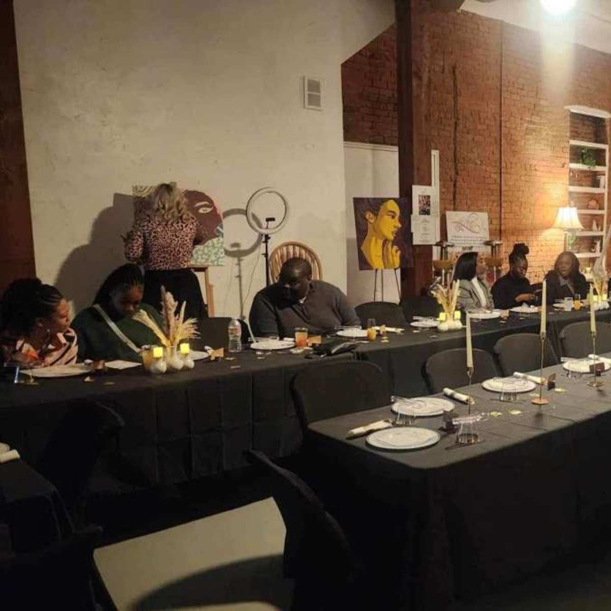 A long table is set for a formal dinner in a space with exposed brick walls, while guests are seated, engaged in conversation.