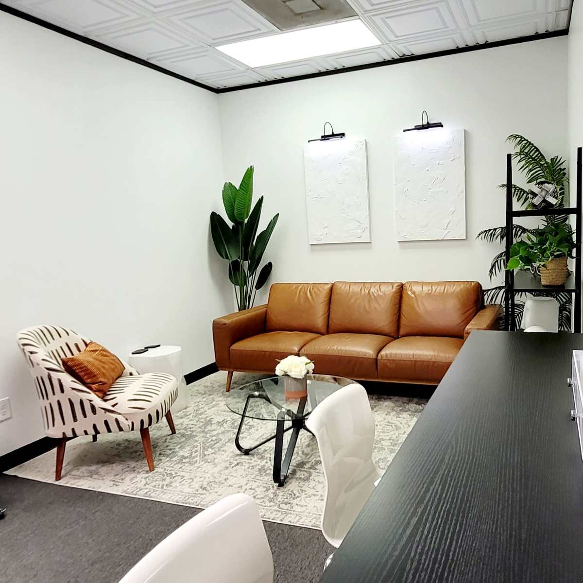 A modern office space featuring a brown leather sofa, a patterned chair, a glass coffee table, and potted plants, with white walls adorned by artwork and a mirror.