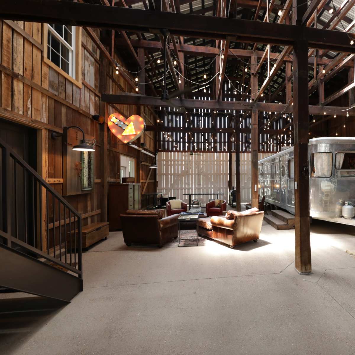 A spacious barn interior featuring an Airstream trailer, a seating area with two leather chairs, and a dining table surrounded by white chairs.