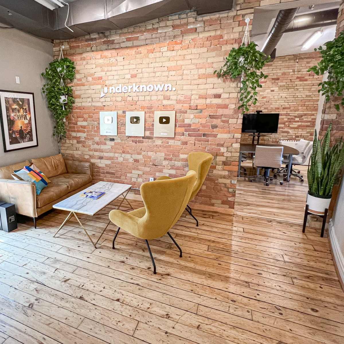 A modern office space with a leather couch, yellow chairs, a coffee table, and a wall decorated with hanging plants and plaques.