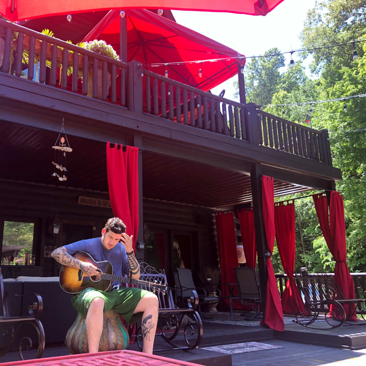 A man plays an acoustic guitar while sitting on a stone in front of a deck with red curtains and an umbrella.